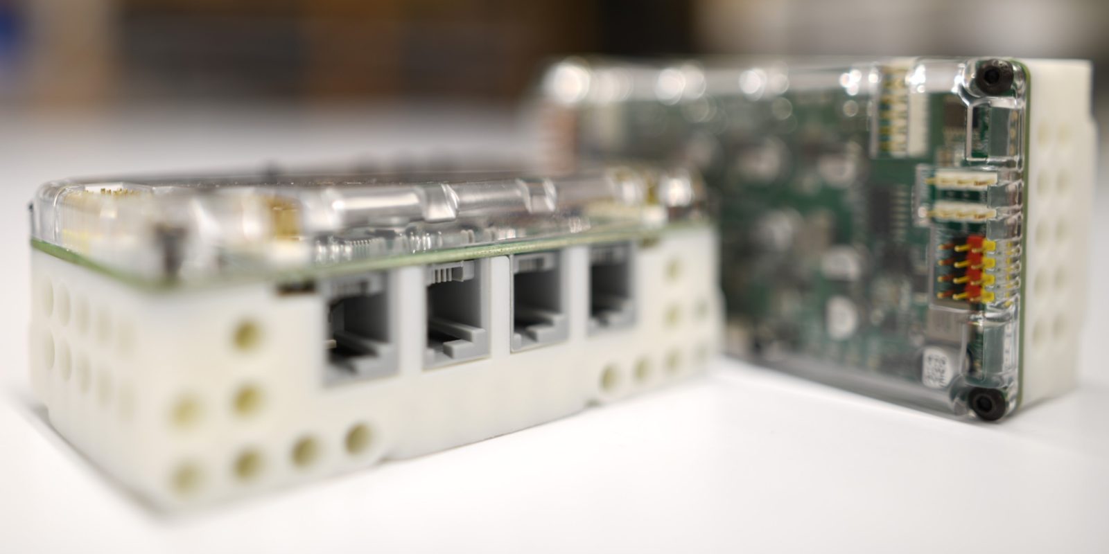 Two EVN ALPHA controllers on a white desk: the front one shows its four RJ12 motor ports in a LEGO-compatible white housing, the one behind it shows the green circuit board through its clear cover.